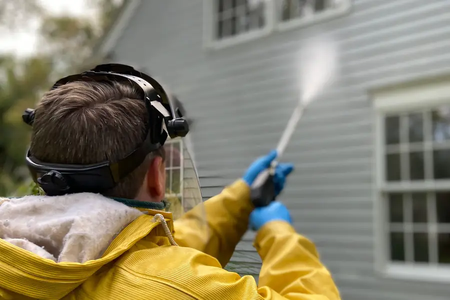 Worker in rain gear and a face shield pressure-washing house siding